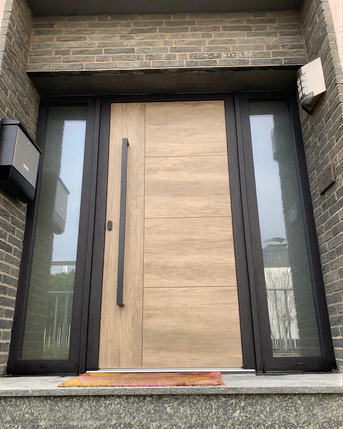 Haustür in heller Holzoptik mit schwarzem Stangengriff und zwei schmalen Glas-Seitenteilen in dunklem Klinker
