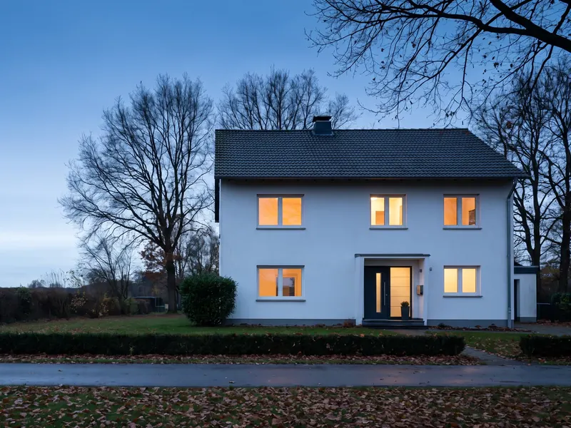 Ein weißes Einfamilienhaus in der Abenddämmerung, in den Fenstern brennt warmes Licht